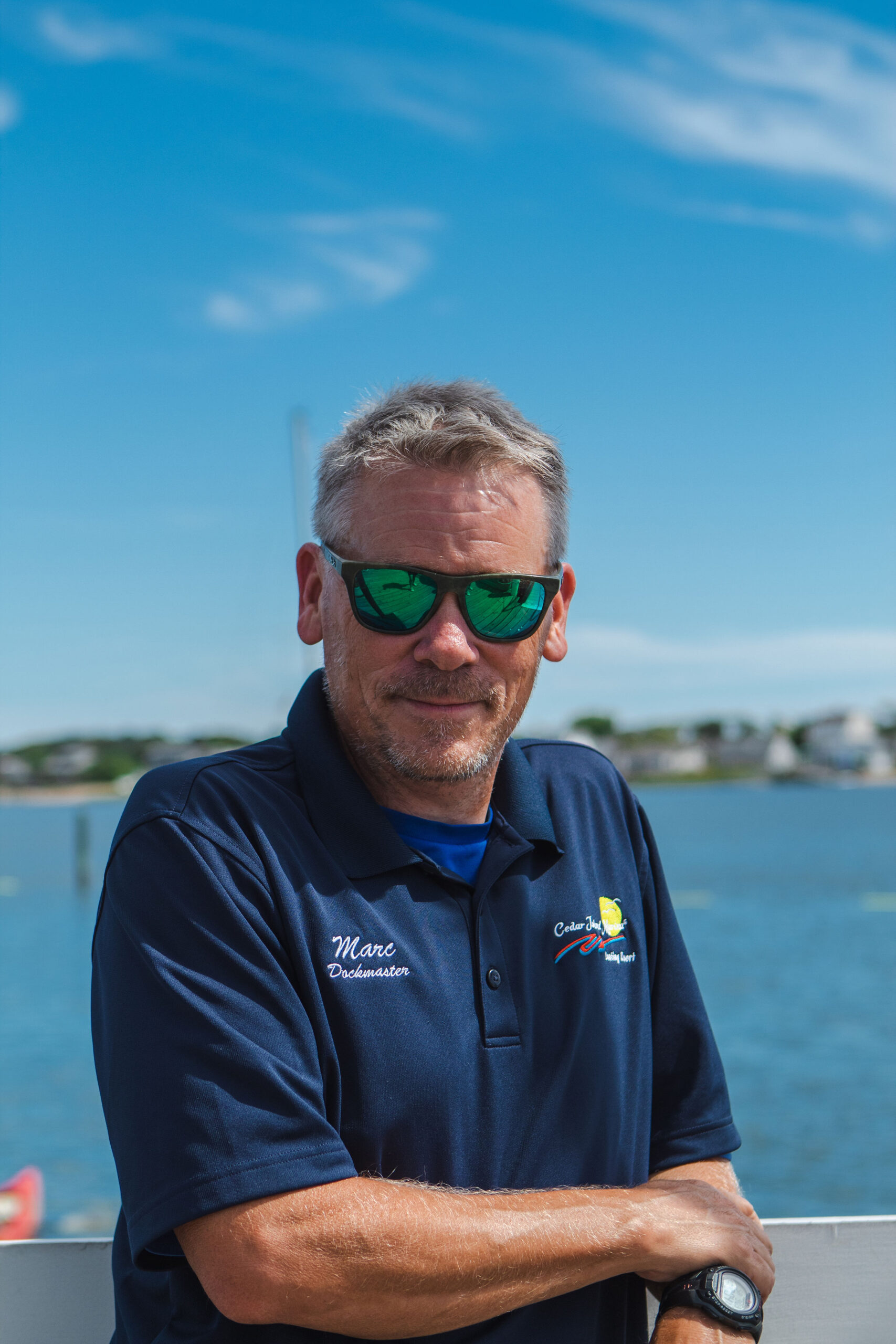 Man wearing a navy polo and sunglasses stands by a railing along a blue harbor, arms crossed in a sunny outdoor setting.